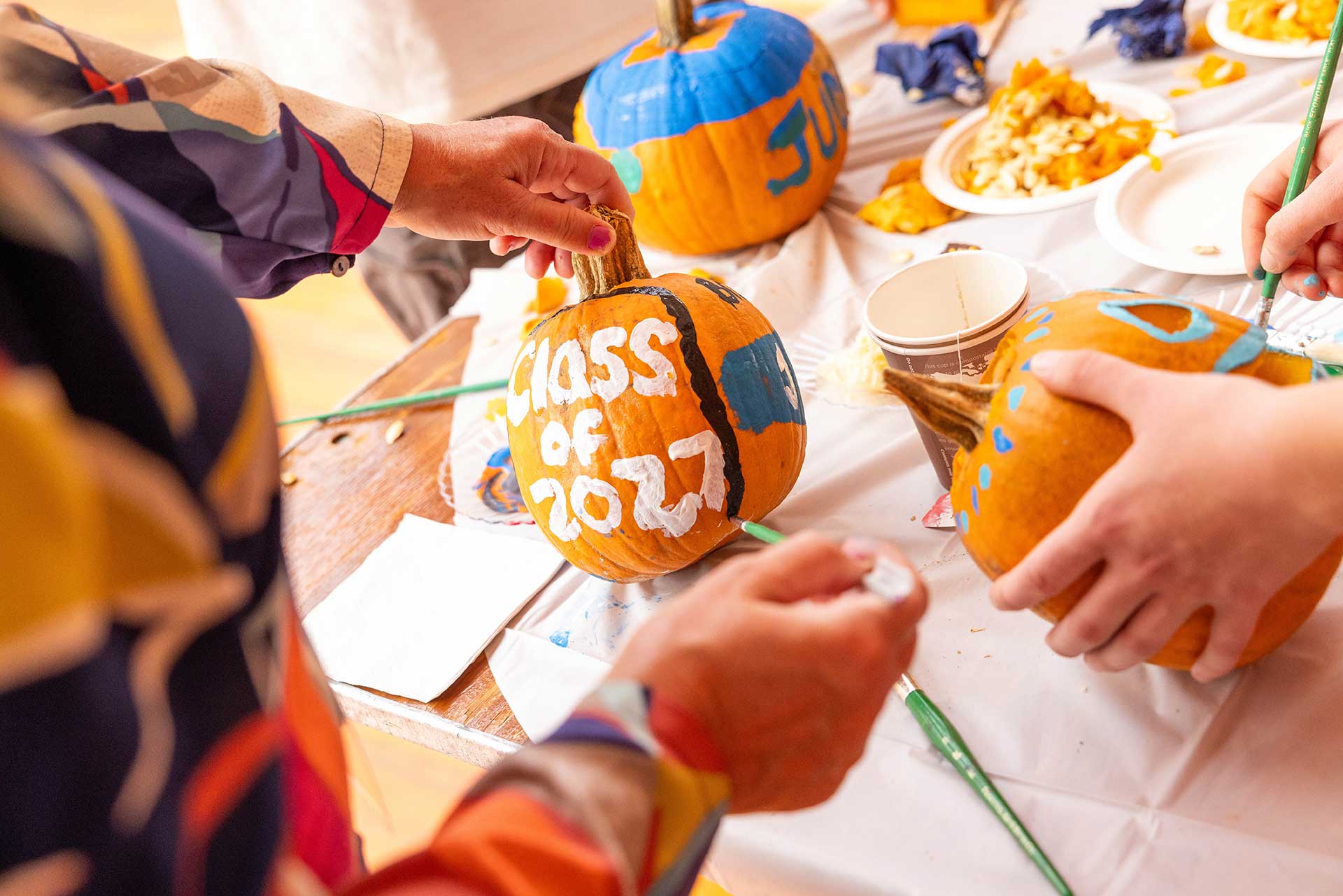 People paint pumpkins. One pumpkin says Class of 2027 in white paint