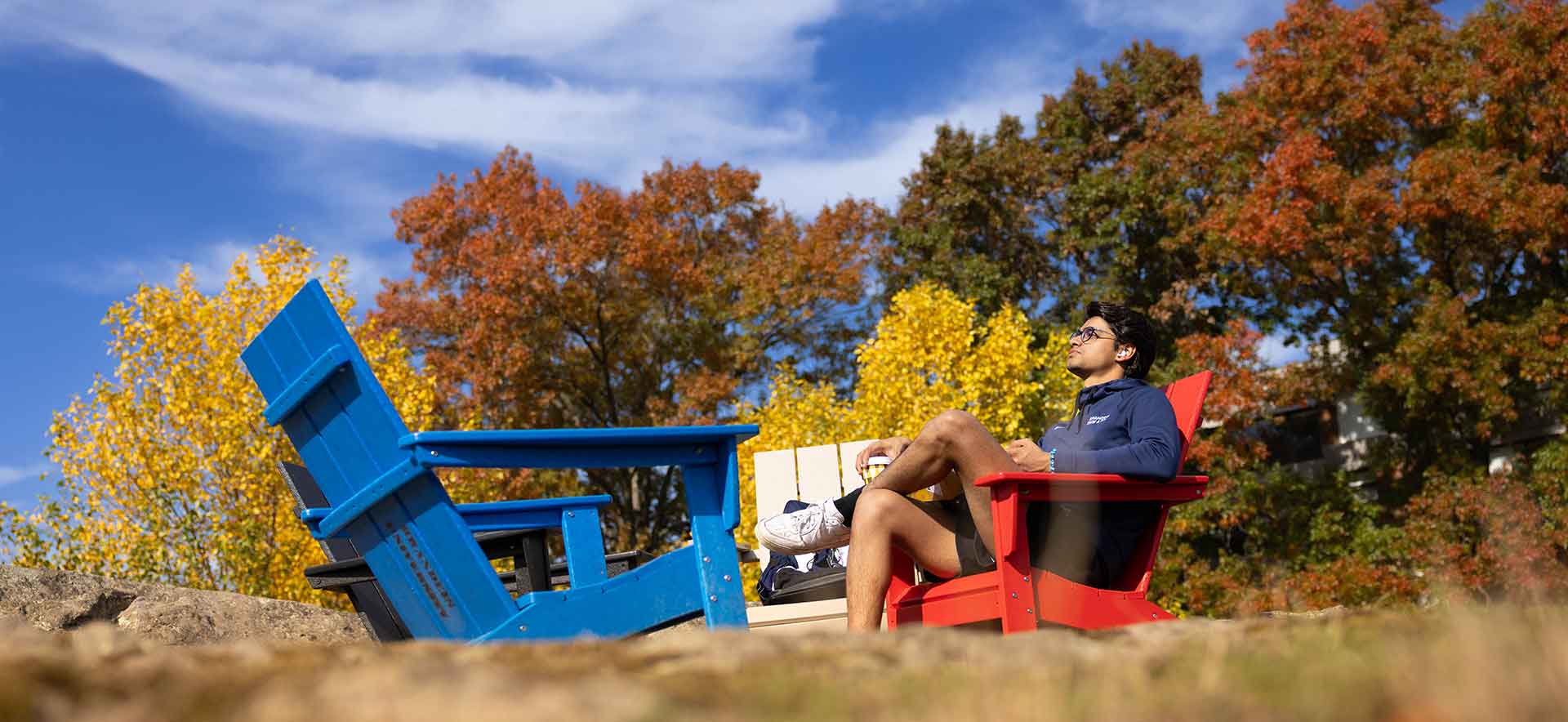 A person sits in a red Adirondack chair. Two empty chairs, blue and white, are nearby.