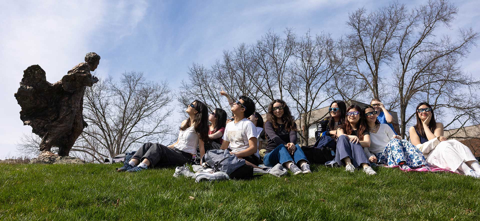 A group of students sit on the grass wearing eclipse viewing glasses and looking at the sky