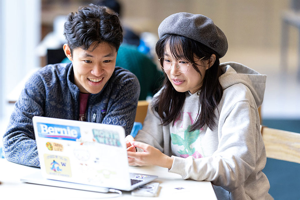 Two students sit together at a table smiling and looking at a laptop.