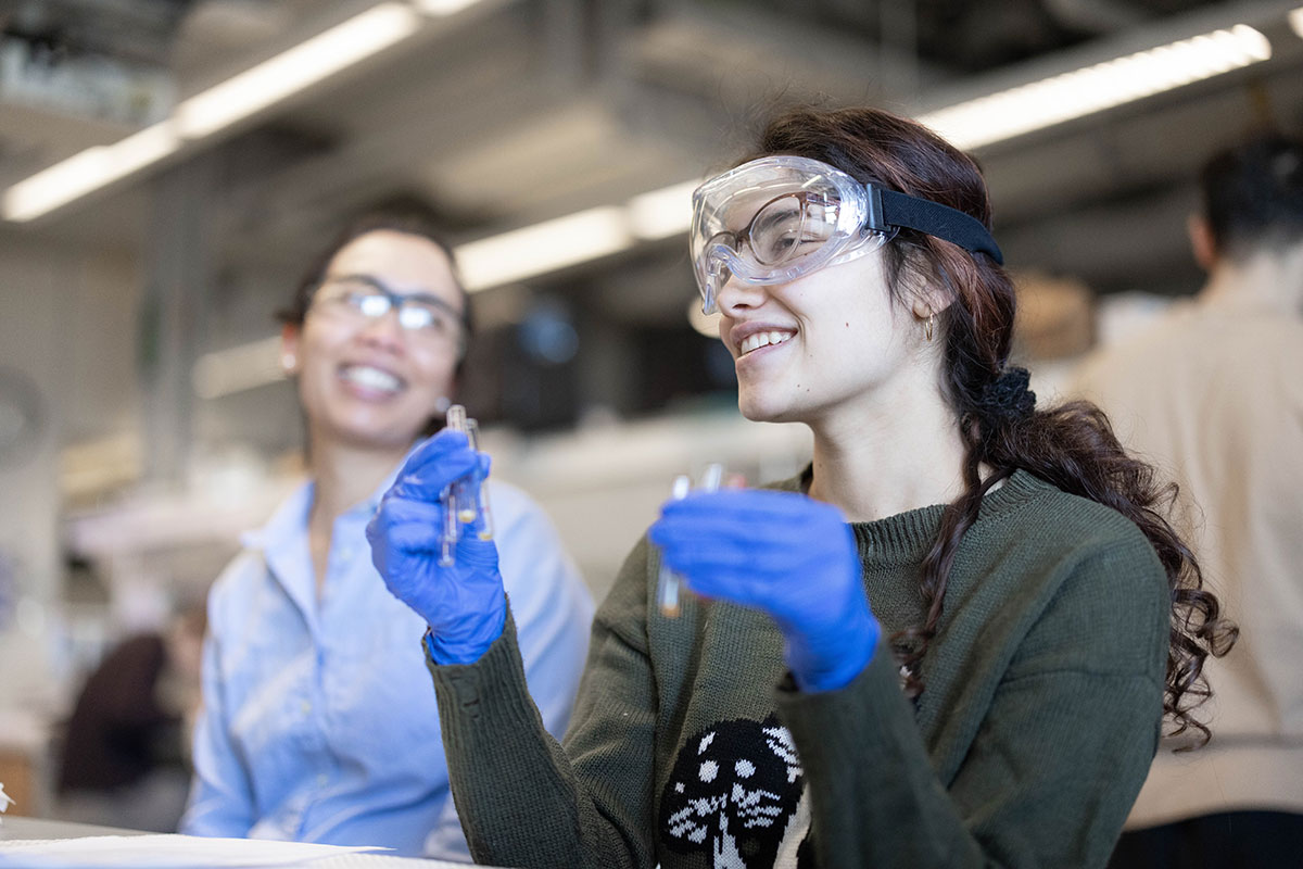 A student smiles in a lab holding a beaker and wearing protective goggles.