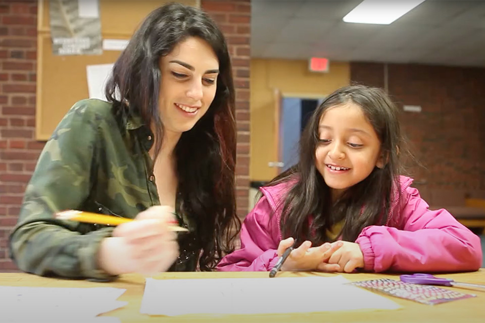 Brandeis student sitting at table with young girl. Both are holding pencils and looking down at a sheet of paper, smiling.