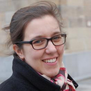 A portrait photo of Elisabeth Gallas wearing glasses and a black coat with a scarf, the white brick walls are in the background.