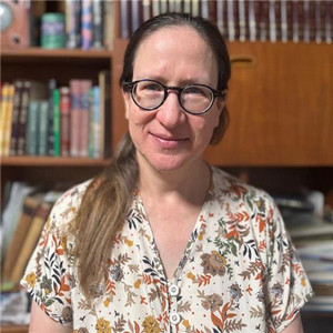 A portrait photo of Havi Ben-Sasson Dreifuss wearing glasses and a shirt with bookshelves in the background