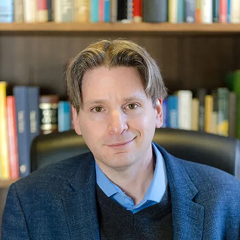 Elias Sacks headshot seated at a desk with books in a bookcase in the background. 