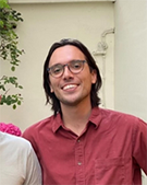 The portrait photo of Trevor Wilson wearing a burgundy shirt and glasses
