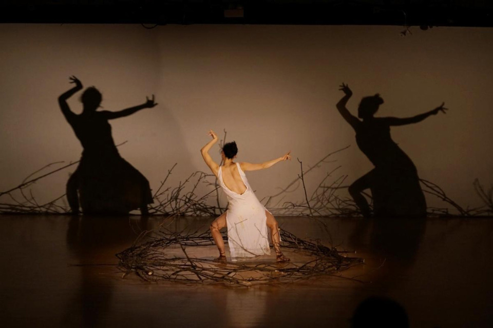 Dancer in a white dress, surrounded by a circle of twigs, dances on a stage with back to the audience.