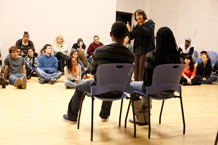 Two students seated in chairs with other students seated on the floor in a theater class with Adrianne Krstansky