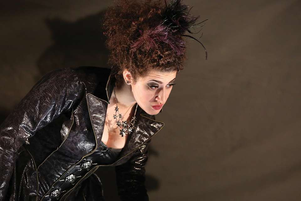 A woman in a black costume with big hair and heavy jewelry looks disgruntled.