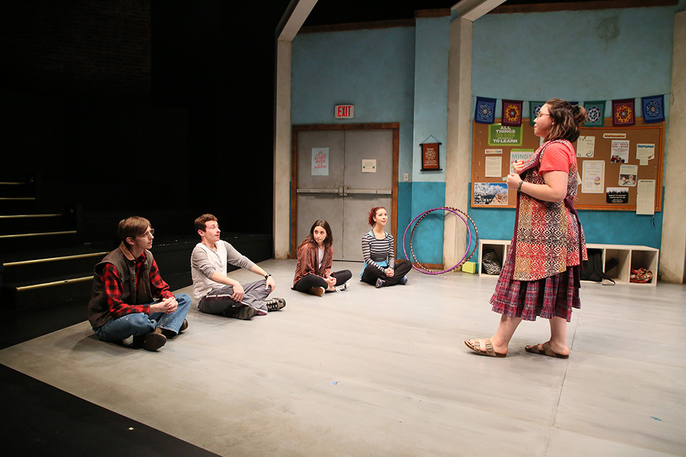 A group of five people, four seated and one standing on a stage with mirrors and cubbies