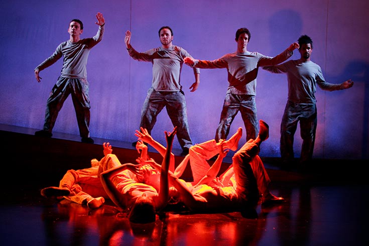 Four actors standing on stage with outstretched arms. In front of them, actors laying on their backs with arms and feet up in the air.