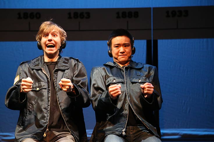 Two male actors seated, holding up their hands as if they were clutching a steering wheel.