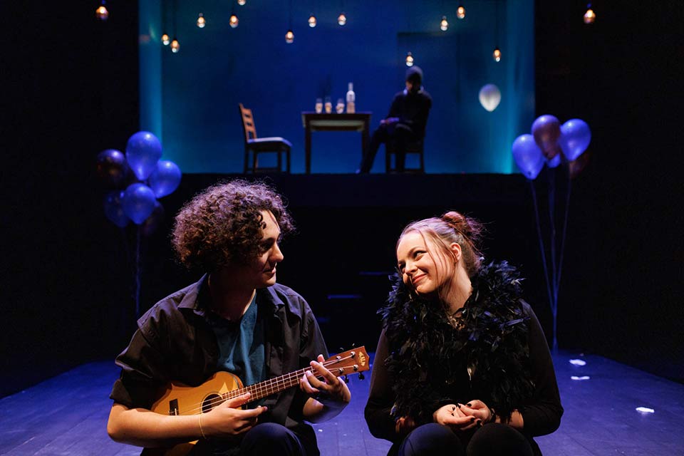 Two actors on stage, one holding a ukulele