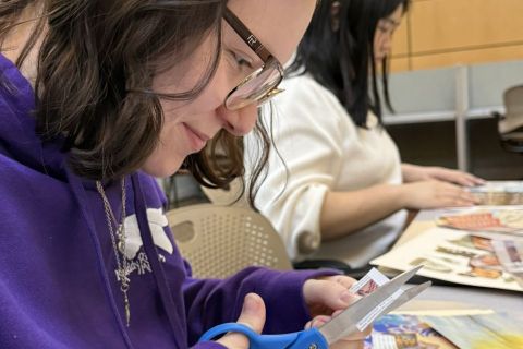 A smiling student surrounded by magazine clippings cuts an image out of a magazine