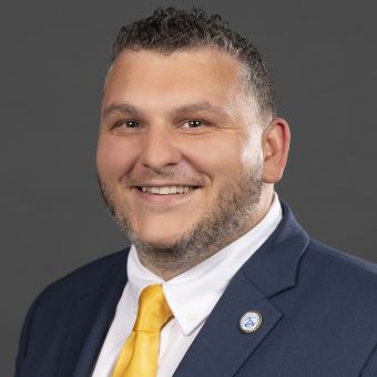 Thomas has short brown hair and wears a blue suit and yellow tie.