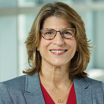Lori wears a grey blouse and dark-rimmed glasses. She has blonde hair.