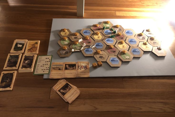 Beside a deck of home-made cards depicting various Greek phrases, a game board shows checkered wooden boxes with Greek lettering across each one.
