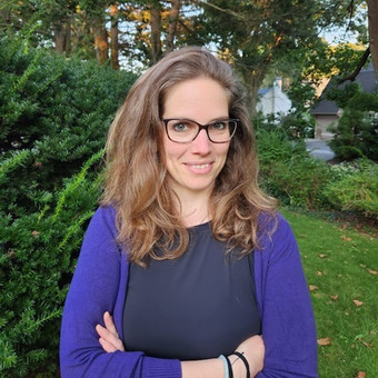 Edith stands in front of a tree. She wears a black shirt with a purple jacket. She has blonde hair and glasses.
