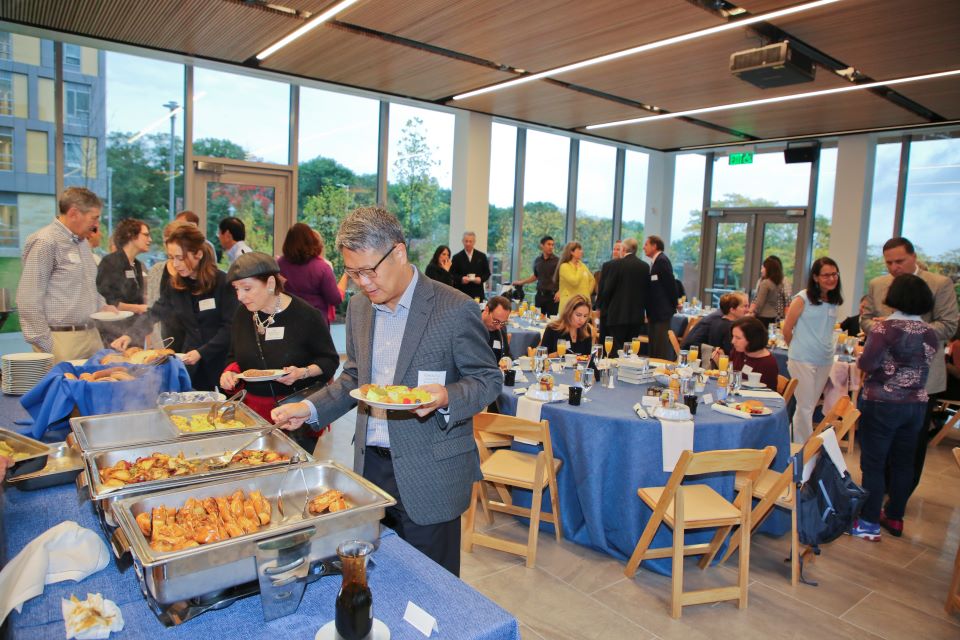 People fill plates at a buffet with the Waltham skyline seen through the windows