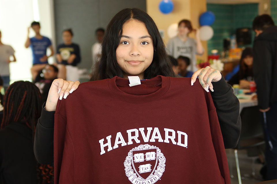 A student smiles and holds up a Harvard sweatshirt.