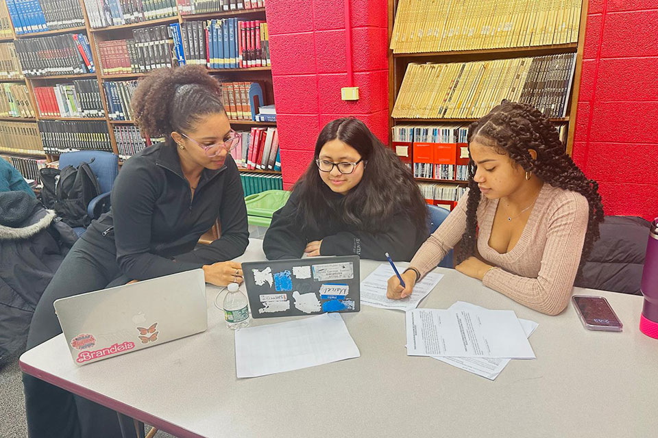Three students sit at a table in a library working on homework.