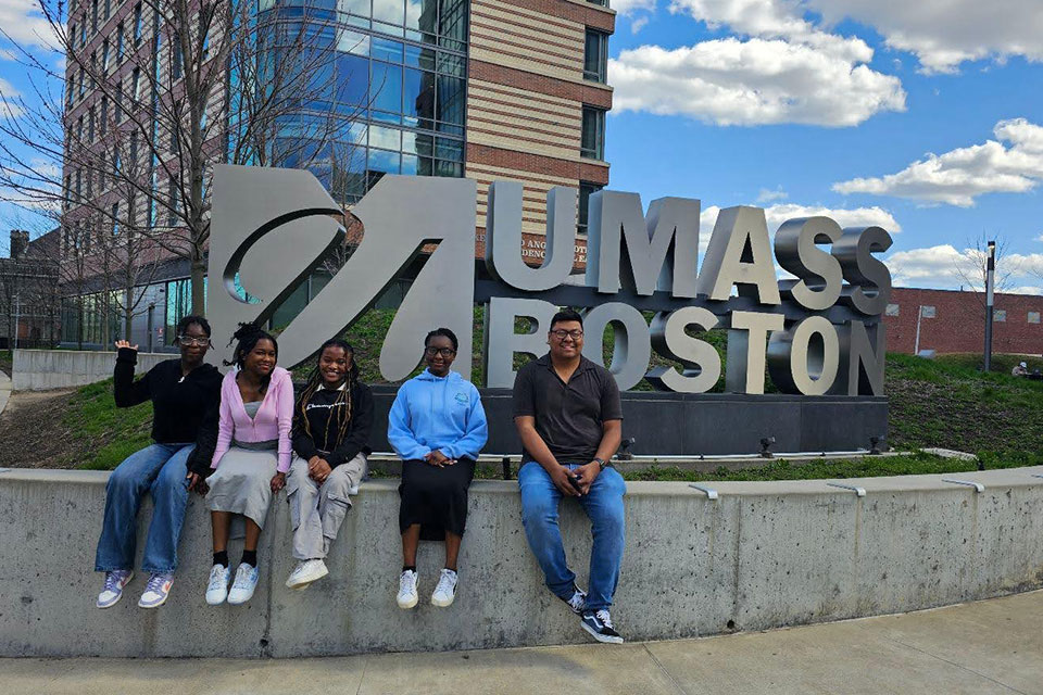 Students sit in front of the UMass Boston sign.