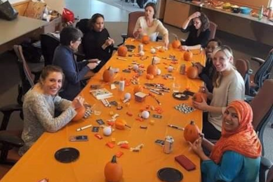 Members sit around a long table with an orange table cloth spangled with pumpkin-decorating bobbles; a couple members hold a paintbrush to their pumpkins and don a smile.