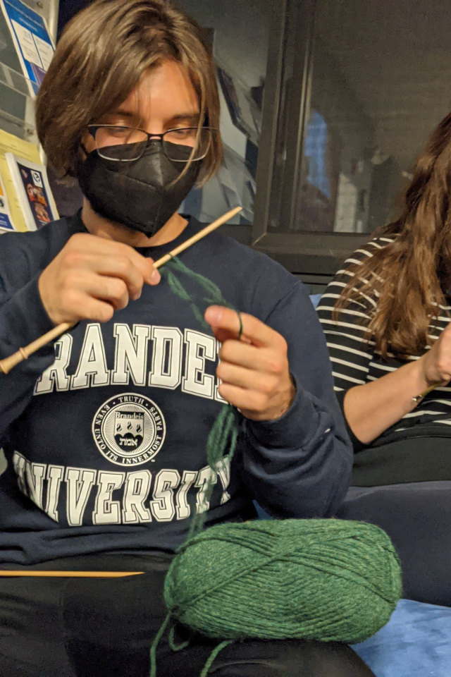 A person in a 'Brandeis' sweater is casting his knitting needle with a long green string of yarn