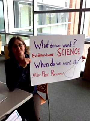 Student holding her poster that says: What do we want? Evidence-based science. When do we want it? After peer review.