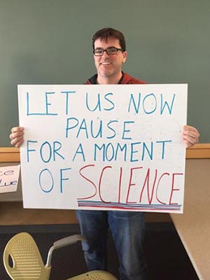 Student holding his poster that says: Let's now pause for a moment of science