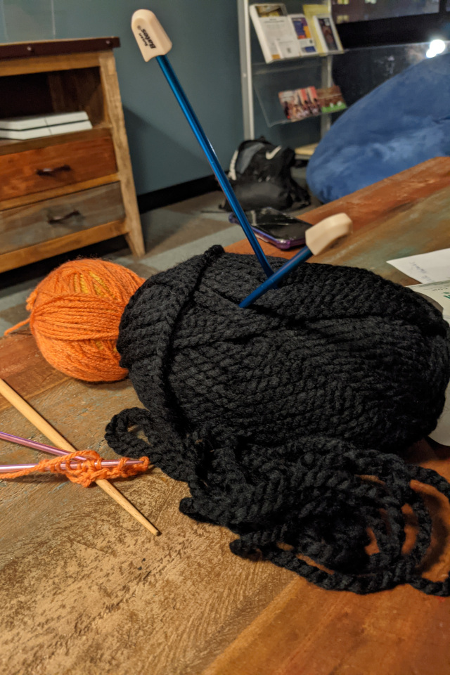 A spool of thick, black yarn sits on a table with knitting needles speared thorugh it.