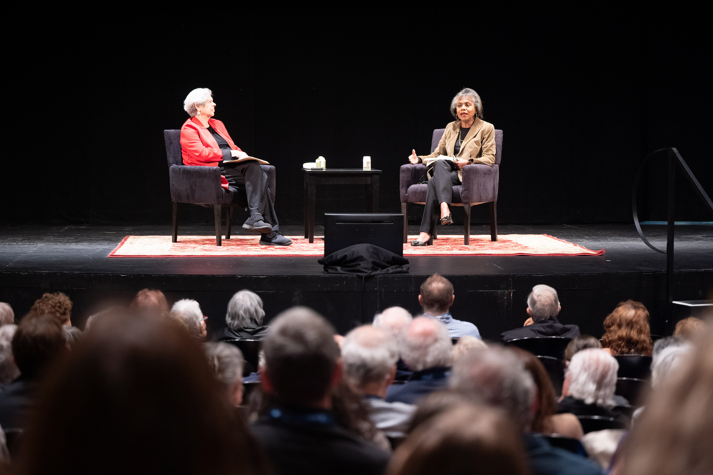 One white and one black woman sitting in chairs on a stage in front of an audience speaking to each other.