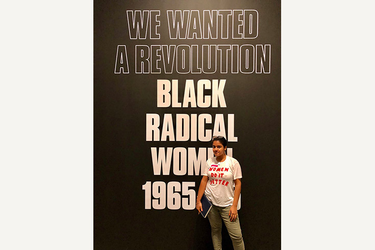 Wearing a shirt that says "Women do it better," Makayla Richards poses in front of an enormous sign that reads, "We Wanted a Revolution: Black Radical Women 1965-85"