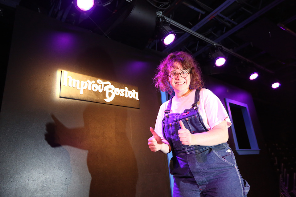 Mina Bond giving two thumbs up on stage at ImprovBoston