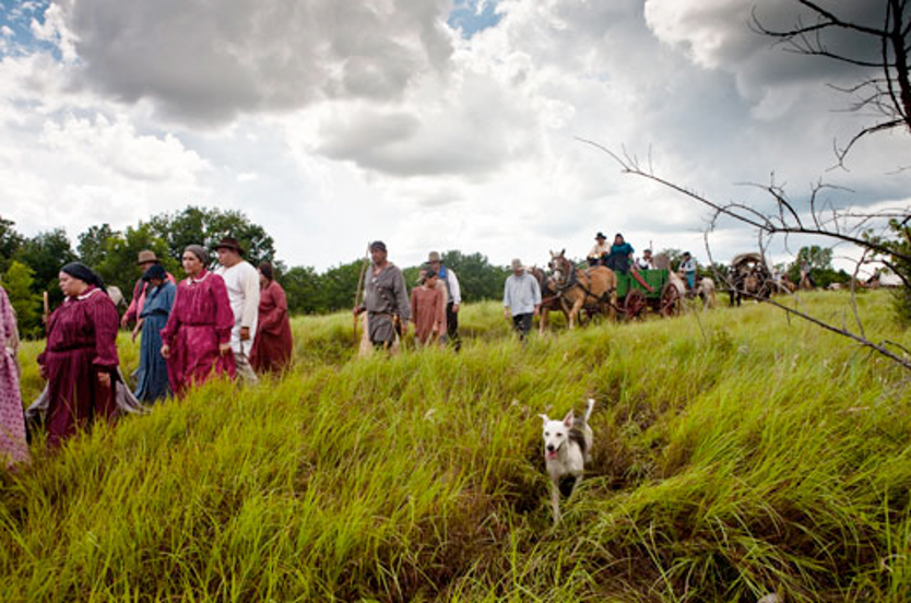 A photograph of Chickasaw men and women traveling in wagons and on foot across a grassy field