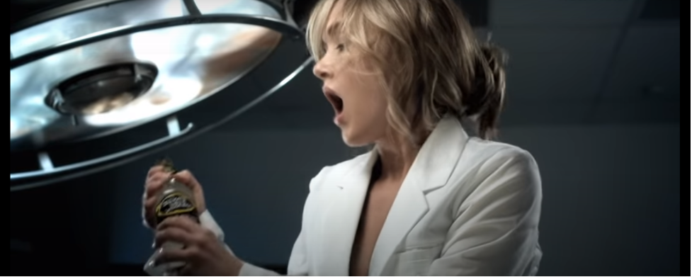 An excited young woman in a white lab coat grips a bottle of Mike's Hard Lemonade