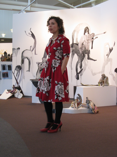 Artist Jessica Riva Cooper at her exhibit “Golem Dybbuk.”  She is pictured here standing in front of a wall that has black and white figures painted directly on the wall.  There are numerous ceramic figures on pedestals of different heights in front of the painted wall. 