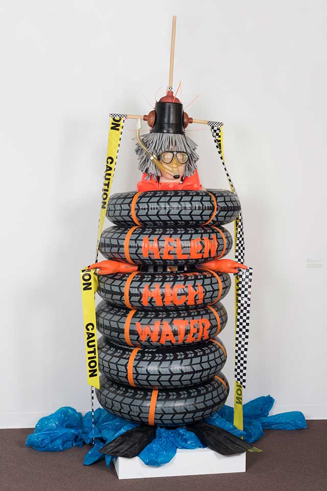Kniznick Gallery Installation: Pat Oleszko, Helen Highwater, 2016. Plastic inner tubes, fabric, plungers, fins, toy cones, dive masks, wig.  This is a sculpture of a woman made of stacked inflatable tires set upon 2 flippers (for feet), orange gloves emerging from the tires on each side for hands, and  a head with a gray plastic wig placed on top. On her head is an upside down bucket, with 3 toilet plungers affixed to the top and both sides.  She is wearing a dive mask which attaches to the toilet plunger.  Yellow construction tape, which says "CAUTION," streams down from the end of the plungers on each side. She is set on a pedestal with blue bunched up plastic resembling water. The words "Helen High Water" are painted in orange on the tires.