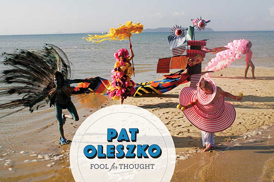 Postcard from Pat Oleszko’s exhibit titled “Fool for Thought” featuring a photo of  "Odds at Sea Bahian Odyssey," a 2009 performance and film, Sacatar Foundation, Itaparica, Brazil. Picture shows several people wearing very brightly colored whimsical costumes, with feathers, balloons, larger than life hats, fanciful creatures with large eyeballs, frolicking on the beach. Text: "Pat Oleszko. Fool for Thought."