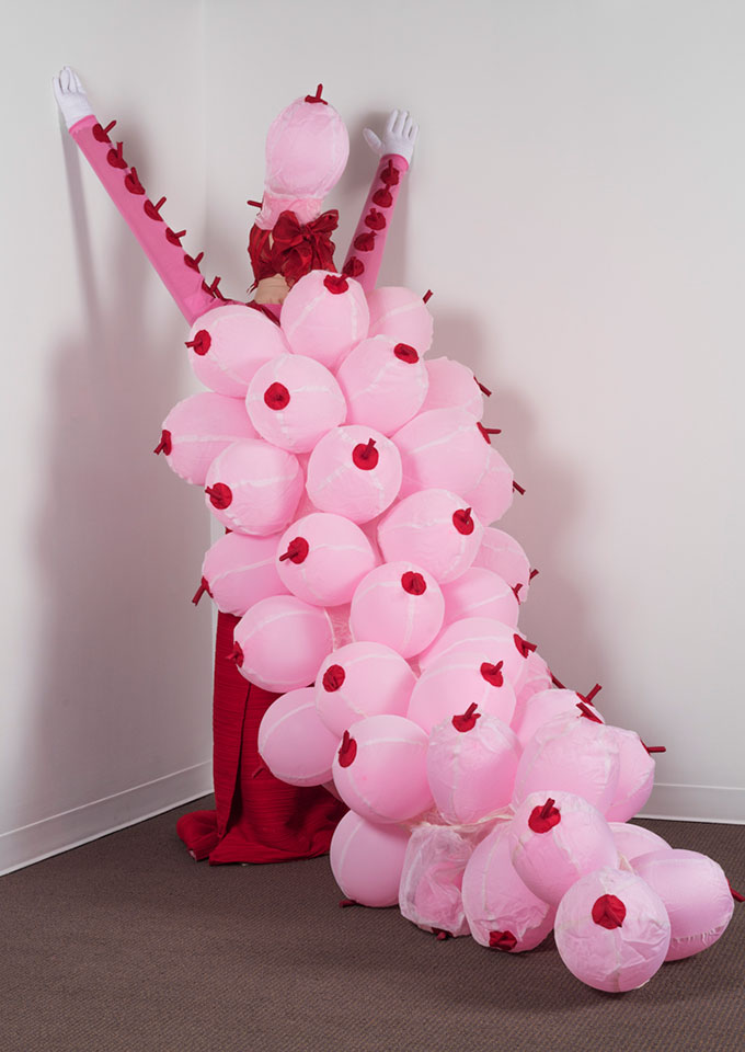 Kniznick Gallery Installation: Pat Oleszko, "Betty Boob," 2007. Balloons, fabric. This sculpture is a woman facing the corner of the room with her hands outstretched above her head, touching the walls.  She is wearing a red floor-length dress and has matching red fabric wrapped around her head with a blow.  The main feature of her attire is a cape made of a cluster of pink balloons, flowing down her back and onto the floor, resembling breasts with red nipples. Her arms have the red “nipples as well, in a row the length of her arms. On her head she wears one of the pink balloons as a hat.