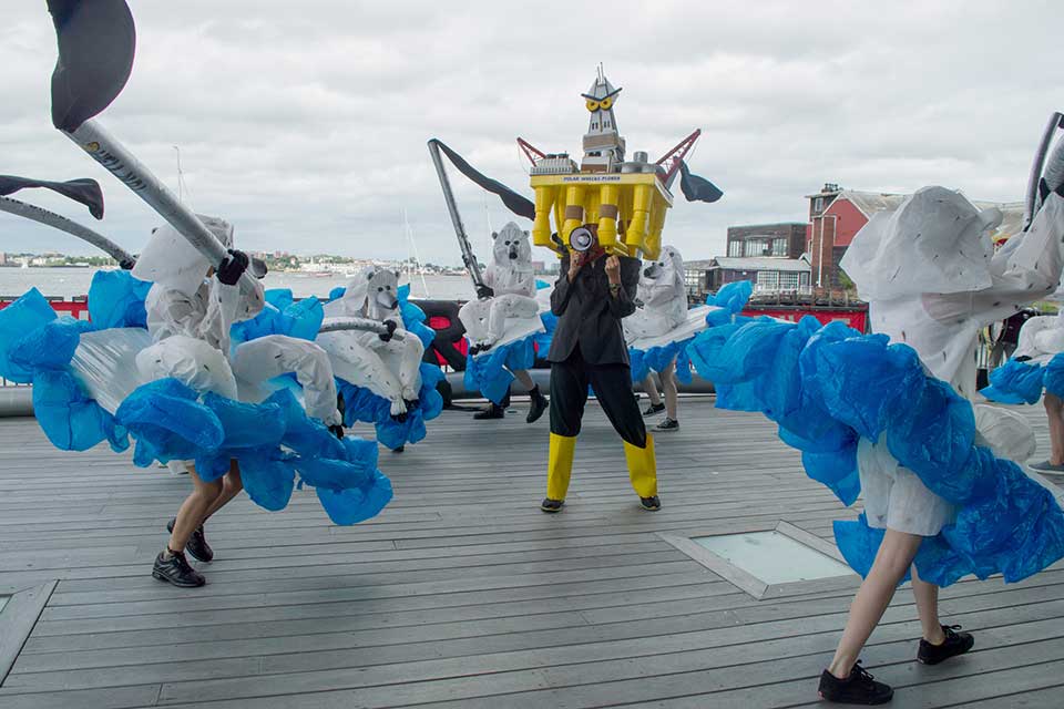 Pat Oleszko, "Hello Folly: the Floes and cons of Arctic Drilling," 2015. Photo of the performance at Institute of Contemporary. Shows people dressed in fanciful costumes of polar bears seated on iceburgs with a blue ruffled "skirt" representing water.  They carry long pipes with black scarves emanating from the ends (perhaps representing oil?).  Each pipe has the words "Hell no". In the center of the circle is someone dressed in a black suit with yellow boots holding a megaphone. On her head is a large headpiece: a yellow oil rig with demonic cartoon eyes. It is labelled "Polar Wrecks-plorer." 