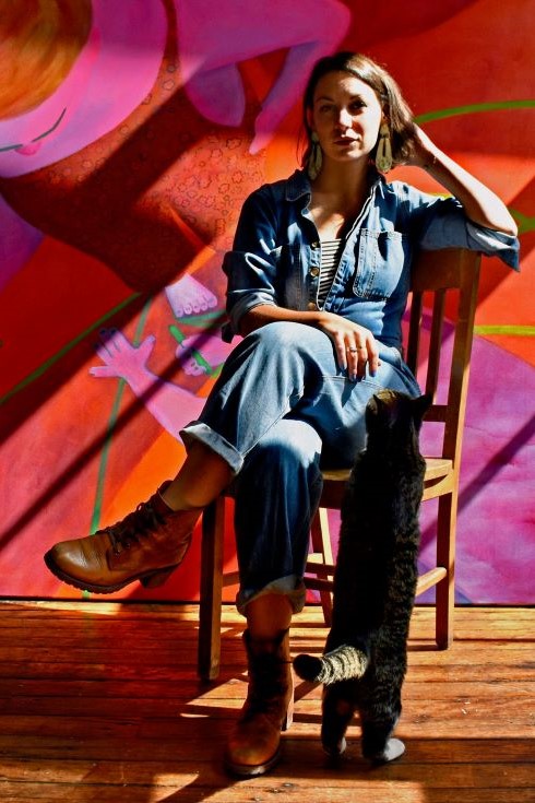 A photographic portrait of artist Genevieve Cohn seated in front of one of her colorful paintings. 