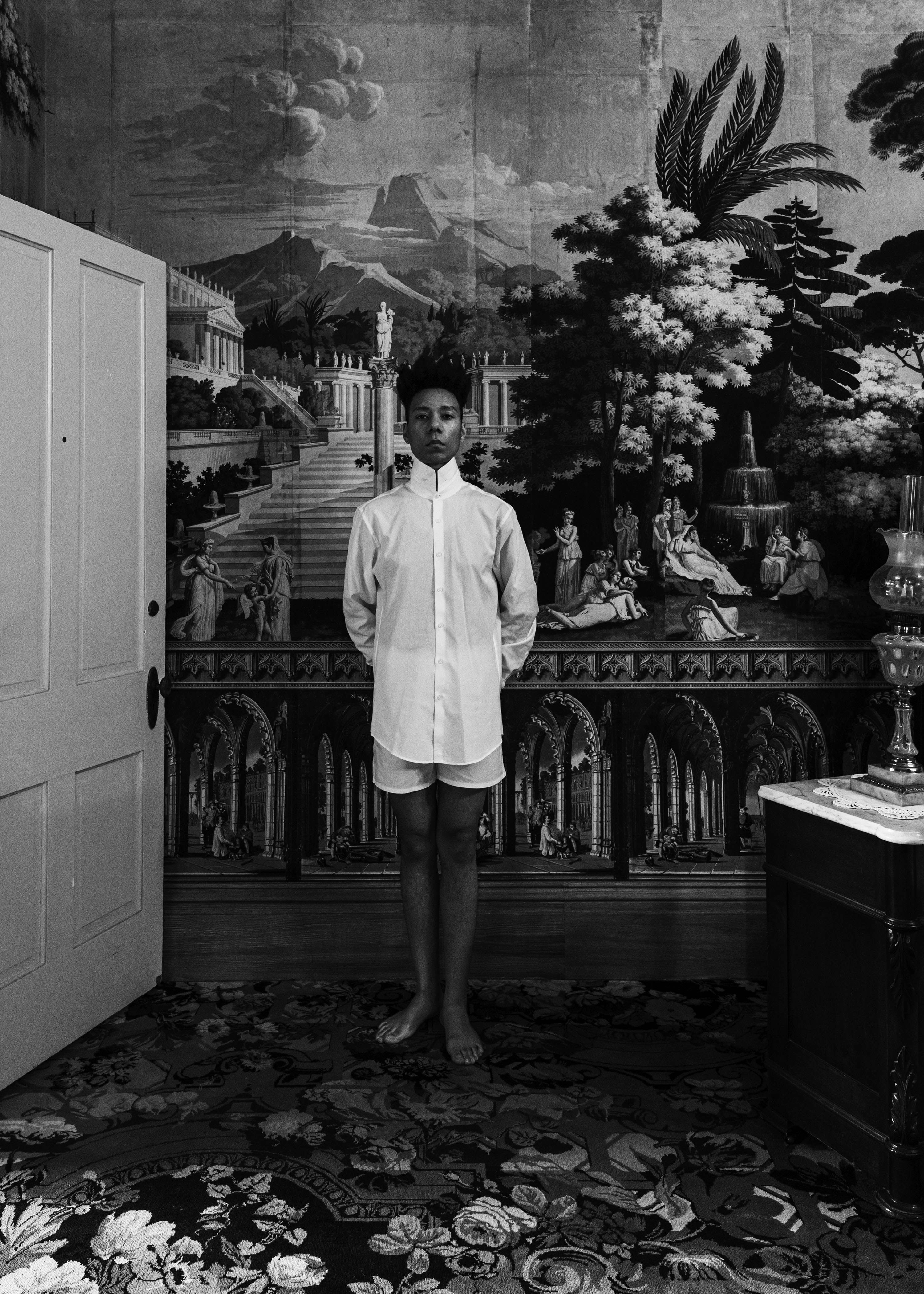 A photograph of a person wearing a white cotton dress shirt positioned in front of an interior wallpapered wall
