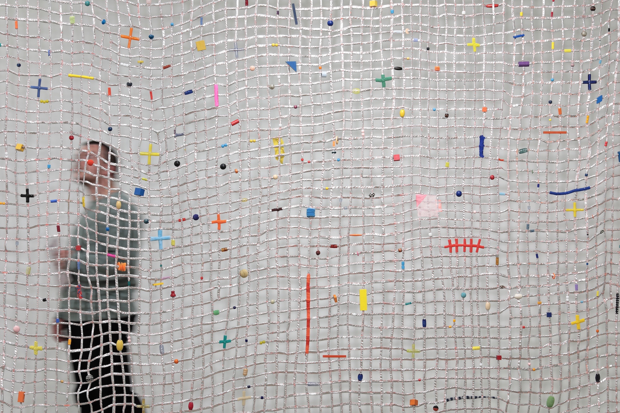 A photograph of Eleanor Anderson's suspended, large-scale, silver, net-like work with colorful adornments and a person visible on its opposite side.