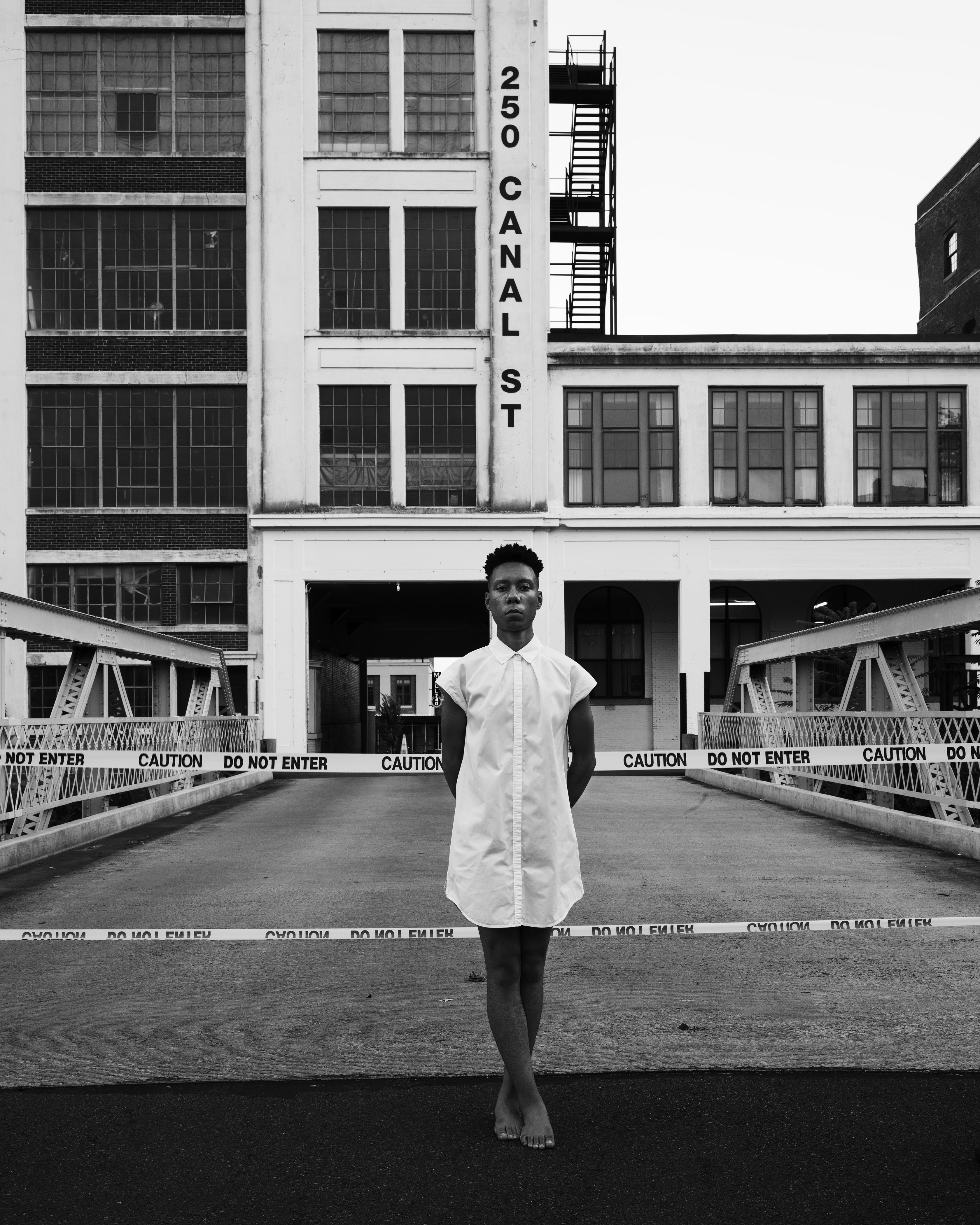 A black-and-white photograph with C. Rose Smith posed in a white dress with legs crossed in front of caution tape with Pacific Mills, Lawrence, MA behind them