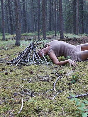 figure on the ground, wearing gray dress, head is covered in a structure made of sticks