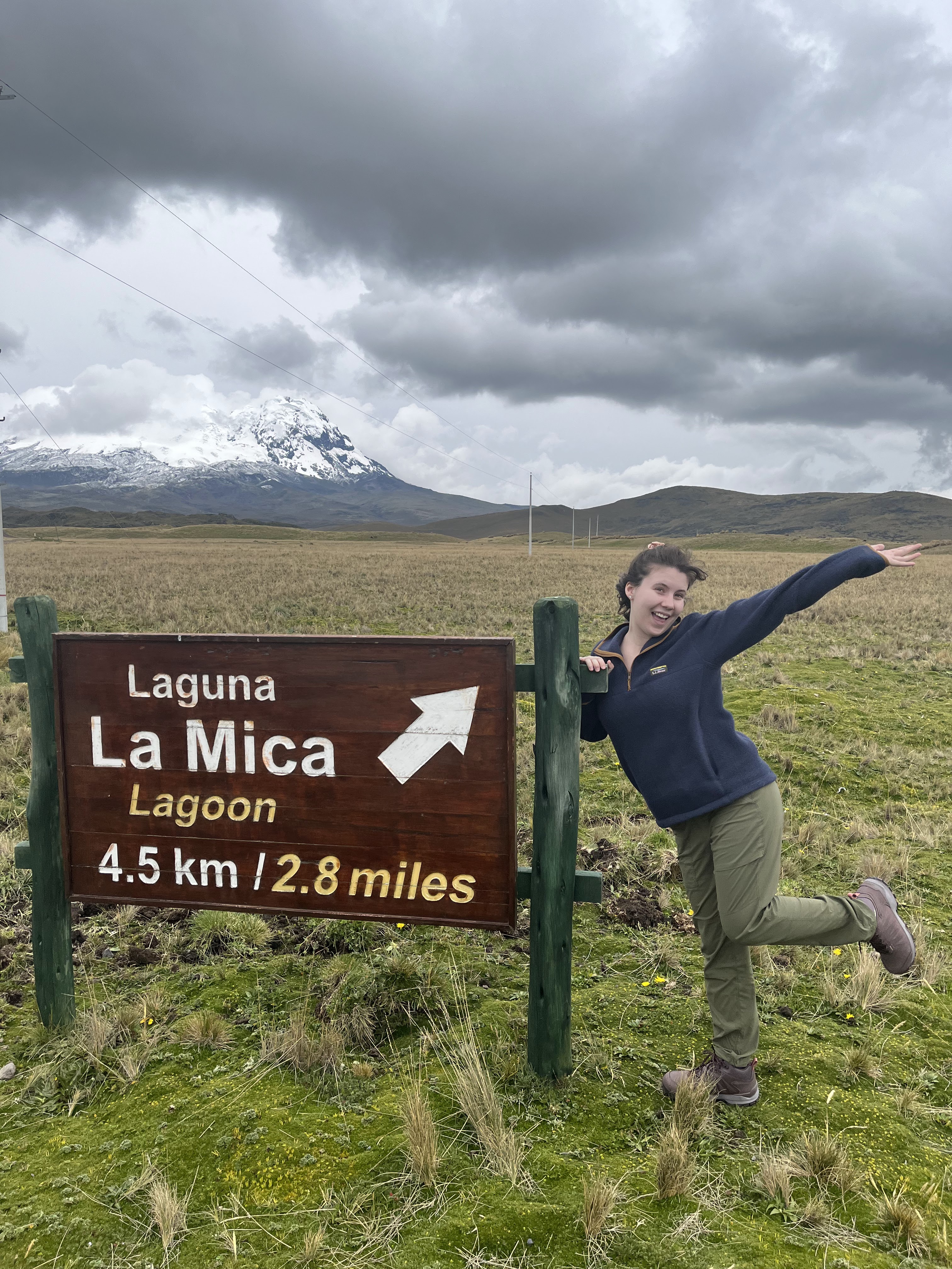 Emily stands on one foot with her arm outstretched against a sign that points toward the lagoon, with rolling hills behind her.