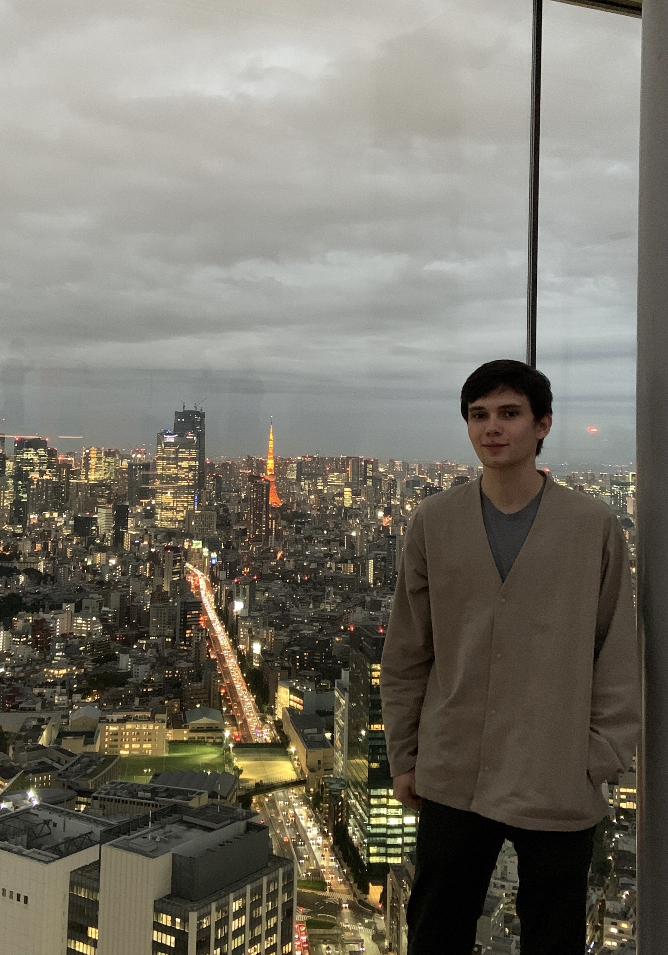 Eben stands smiling against a glass wall; beyond, the cityscape of Tokyo shines at sundown.