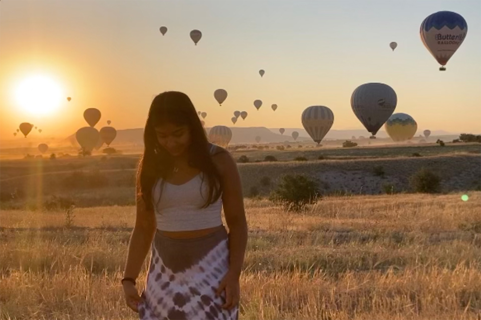 A student walking in the foreground with hot air balloons and a sunset behind her