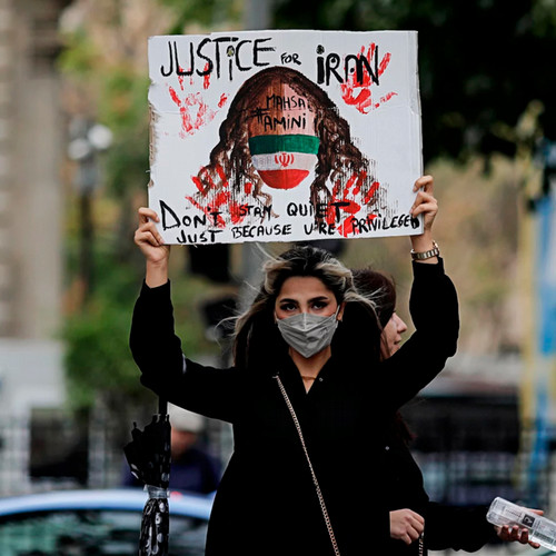 An Iranian woman dressed in black without a hijab and wearing a paper face mask holds up a sign with red handprints and the text, "Justice for Iran, Don't Stay Quiet Just Because U're Privileged" with a woman's face and hair in the center.